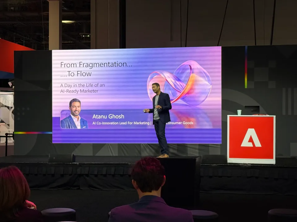 A speaker on the keynote stage in front of a slide reading 'From Fragmentation... To Flow', a red Adobe cube beside him.
