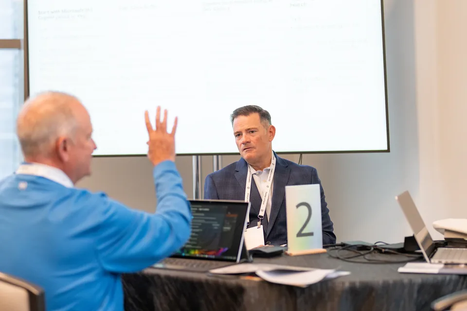 Two attendees talking across a table of laptops at a numbered breakout station.