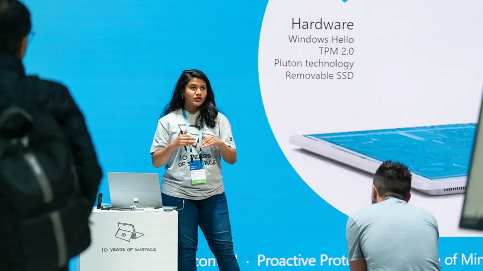A speaker presenting a Surface hardware slide - Windows Hello, TPM 2.0, Pluton, removable SSD - to a seated audience.