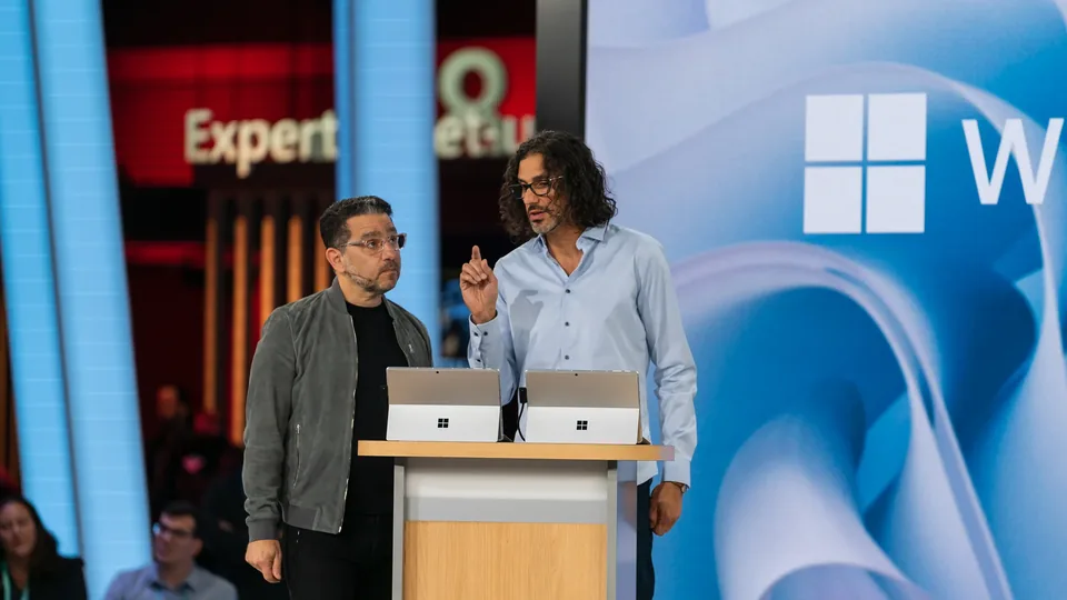 Two presenters at a lectern of Surface devices in front of a large Windows logo.