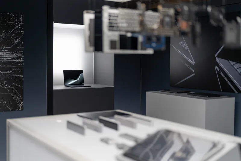 Suspended device panels above a lit display plinth in a darkened product area, laptops arranged on the surface below.