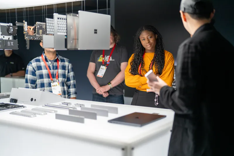 Attendees in lanyards gathered around a lit plinth of Surface devices while a host talks them through the hardware.