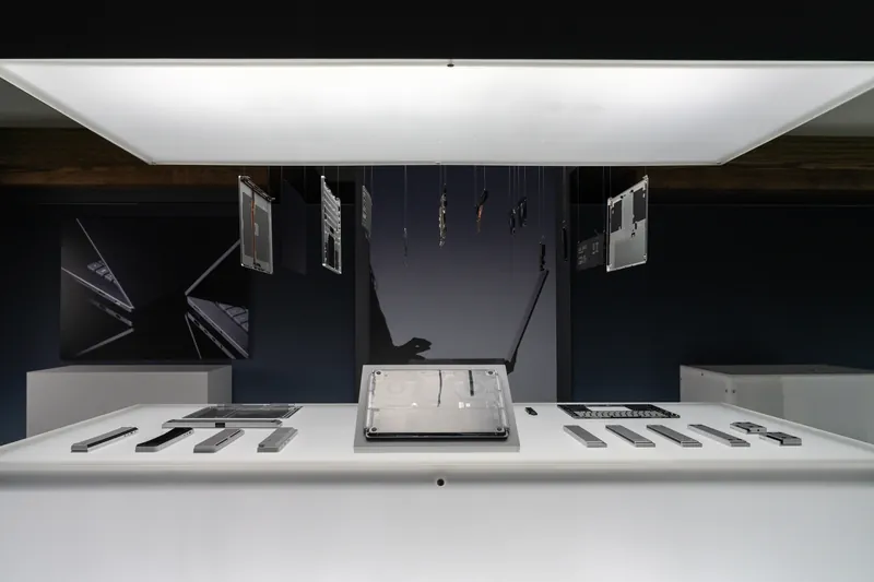 Wide view of the darkened hardware gallery: a broad overhead light panel above a plinth of components, device panels hung on wires.