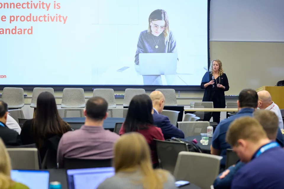 A speaker presenting to a seated audience, the slide behind reading that connectivity is the productivity standard.