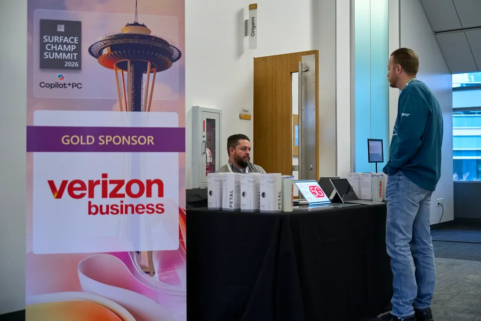 An attendee at the Verizon Business sponsor table, devices laid out along it under a Surface Champ Summit banner.